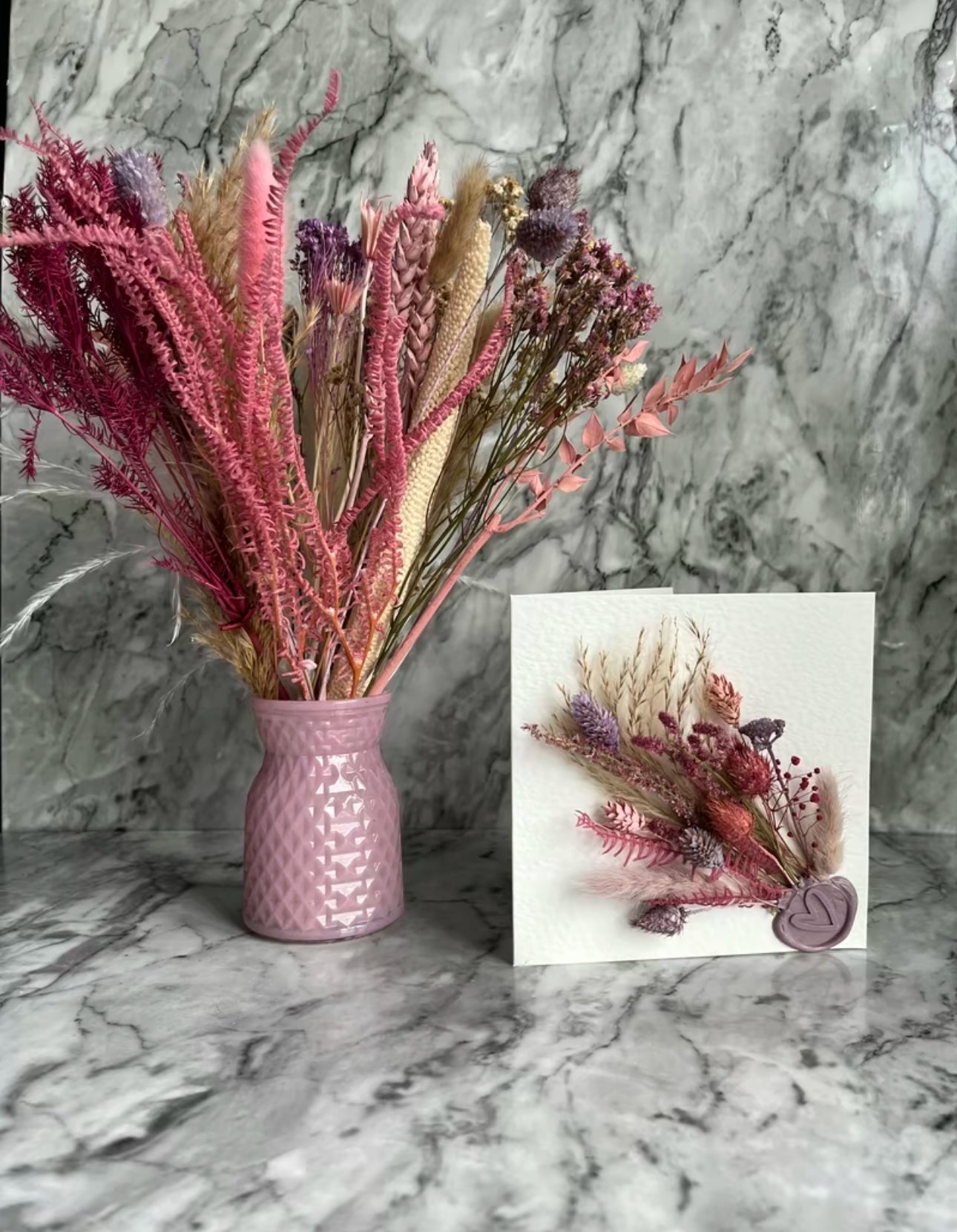 Pink vase with pink dried flowers and a dried flower card on a table