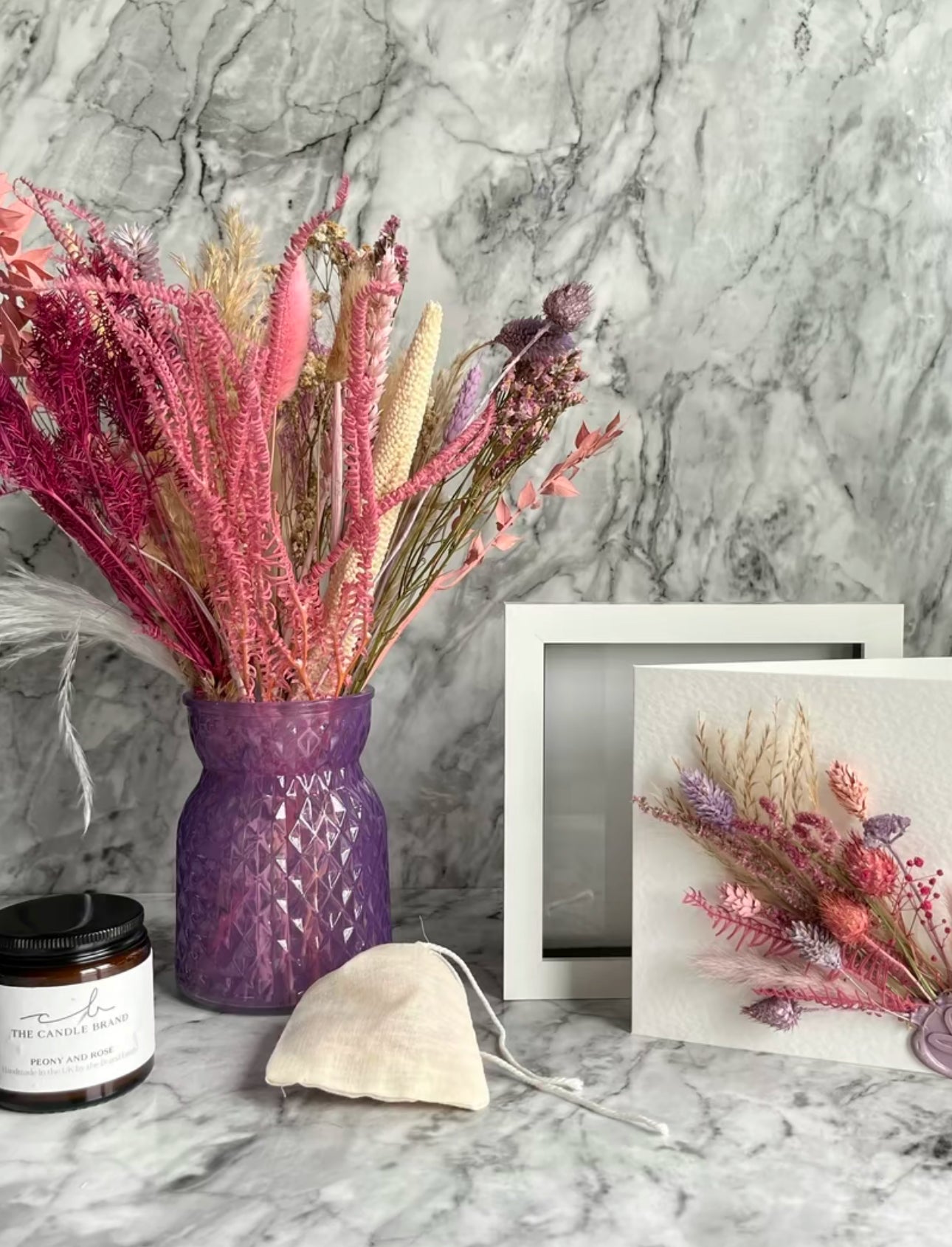 Pink and white dried flowers in a purple vase with a card, candle, frame and bag