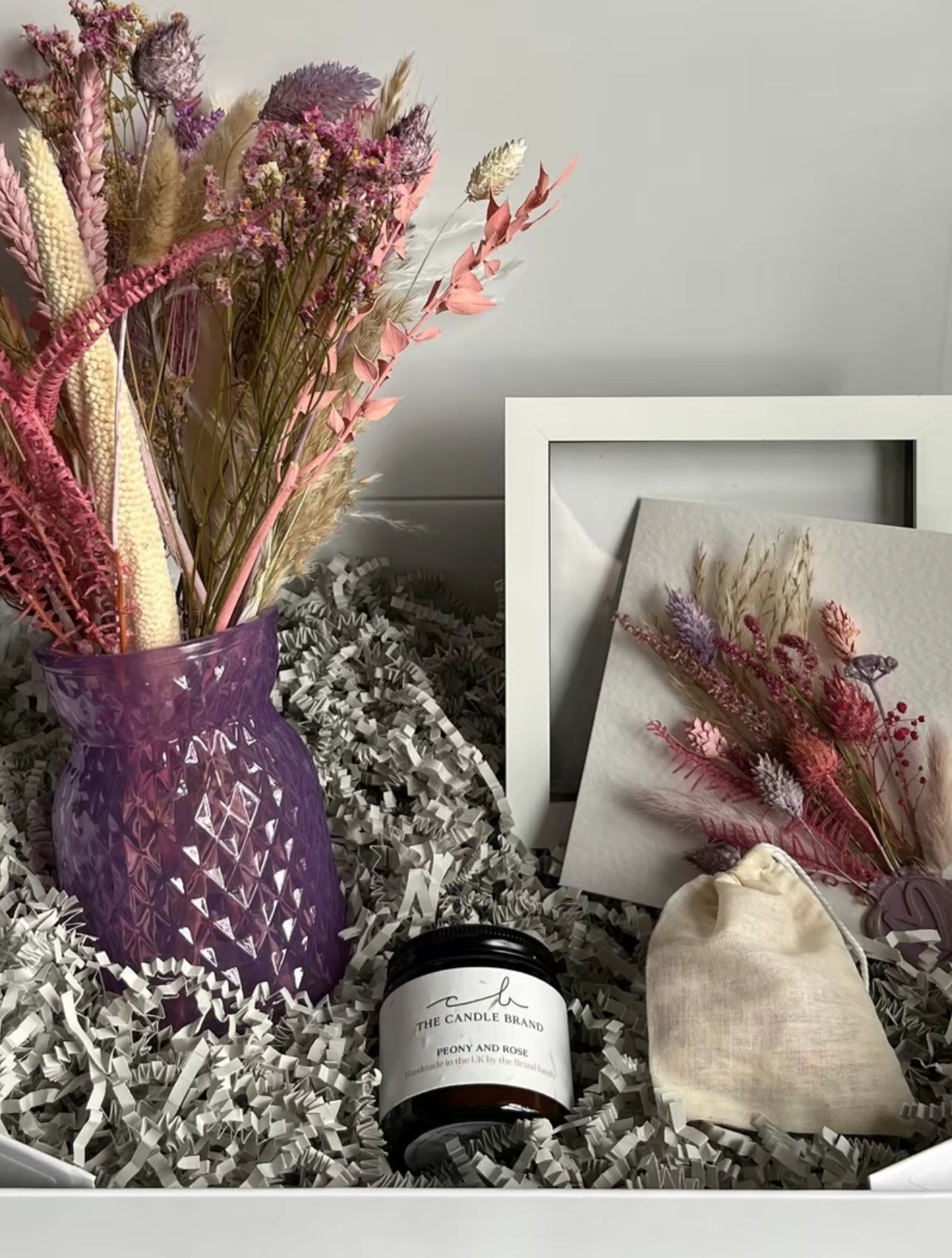 Pink and white dried flowers in a purple vase with a card, candle, frame and bag in a box